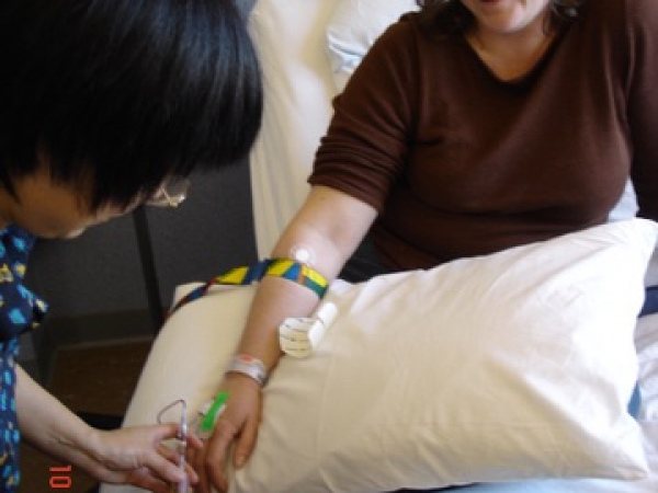 Chandra in hospital bed, her bandaged arm rests on a pillow, as a nurse is checking the arm.